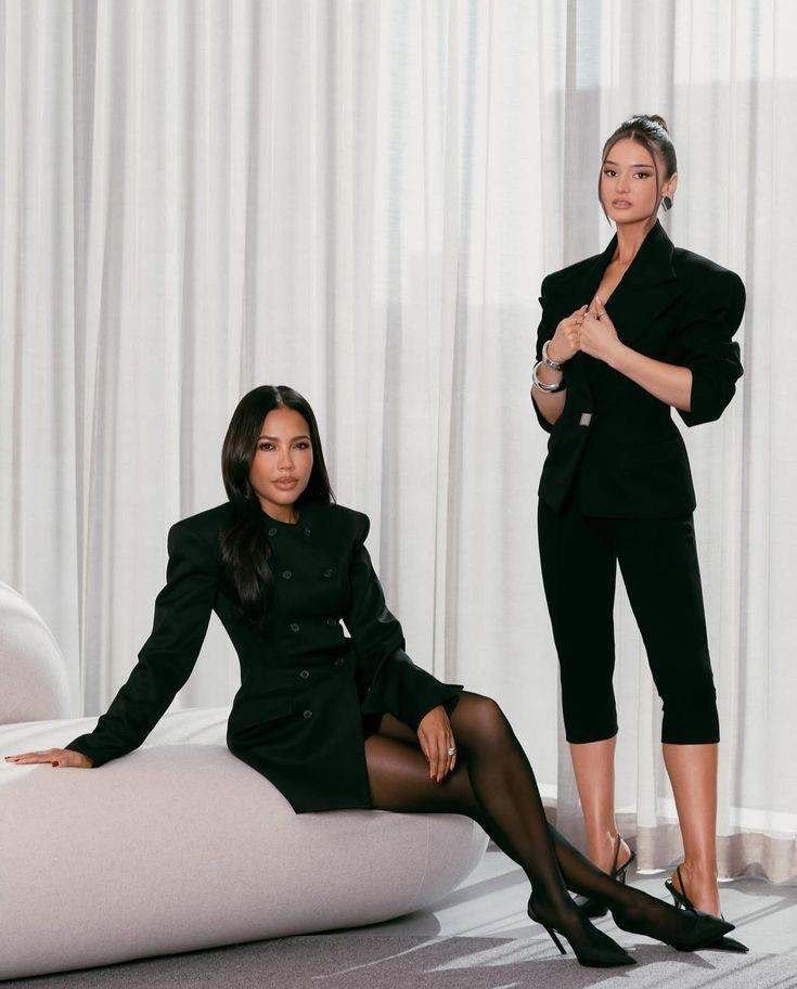 Two women in dark tailoring seated in a neutral studio
